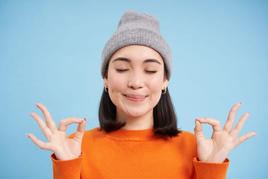 Sounds mind in healthy body. Smiling calm and relaxed asian girl in beanie, shows zen, relaxation gesture, meditating, standing over blue background