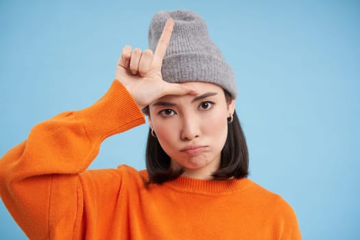 Close up portrait of asian girl student in beanie, shows L letter, loser sign on forehead, stands over blue background