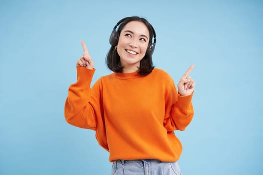 Beautiful korean woman in headphones, dancing and listening music in earphones, standing over blue background