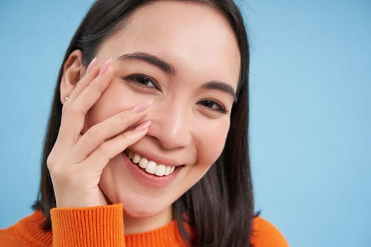 Beauty and skincare. Close up portrait of happy smiling japanese woman, touches her clear, glowing skin, natural healthy face, standing over blue background