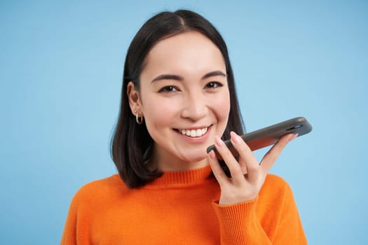 Portrait of asian woman records voice message, talks on speakerphone, holds mobile phone near mouth while speaking, translates her speech on app, blue background