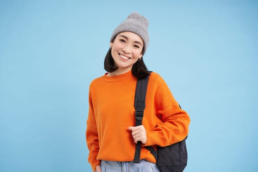 Stylish young asian woman in warm hat, walking with backpack, going somewhere with bag, standing over blue studio background