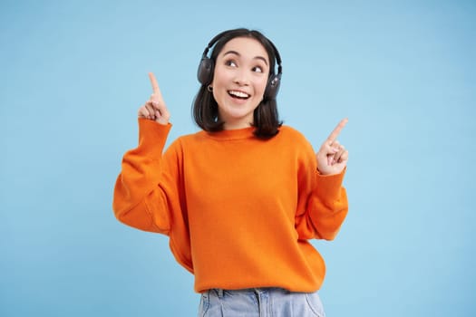 Happy Chinese woman in headphones, listens music, enjoys favourite song in her playlist, stands over blue background