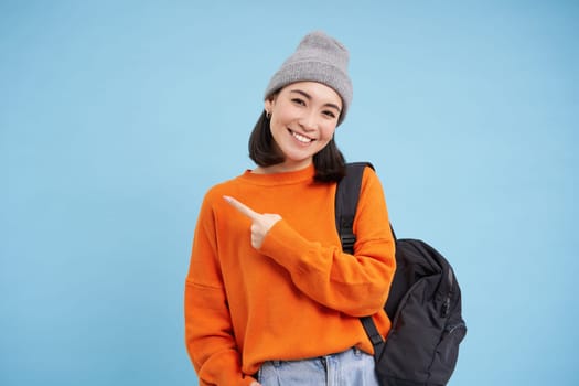 Enthusiastic asian woman, points finger left at banner, shows promo offer, smiles and looks happy, stands in hat with backpack, blue background