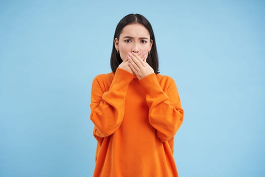 Portrait of shocked korean woman gasps, closes her mouth with hands and look speechless at camera, stands over blue background