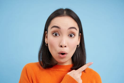 Close up of surprised asian girl, pointing finger right, showing advertisement with curious, intrigued face, standing over blue background