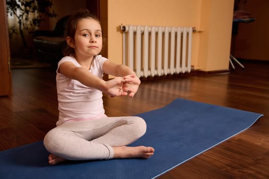 Caucasian lovely kid girl 5 years old, sitting barefoot on yoga mat, stretching arms, looking confidently at camera