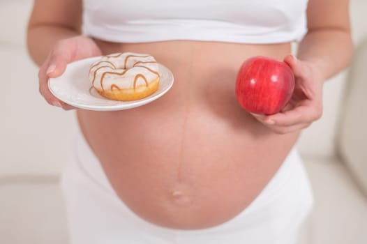 A pregnant woman is holding a red apple and donut. Choice of food.