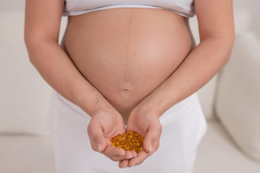 A pregnant woman with a bare belly holds a handful of vitamins. Transparent golden pills. Vitamin D for the expectant mother.