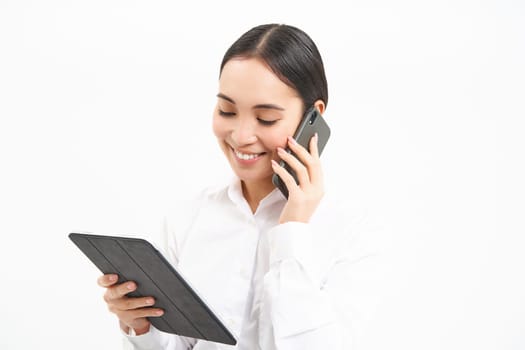 Asian working businesswoman discussing business over the phone, talking on cellphone and looking at digital tablet, standing over white background