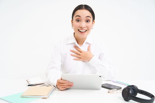Image of asian businesswoman holds tablet, looks surprised and amazed at camera, hears great news, white background