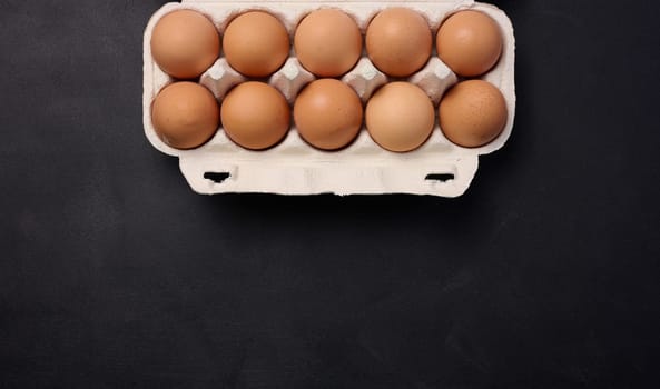 Raw brown chicken eggs in a paper cardboard container on a black background, top view