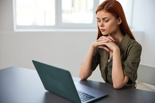a serious, work-weary woman, with a sad face, sits at a laptop with her hands folded near her face