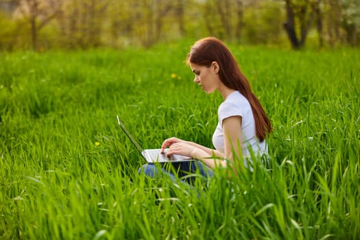 female freelancer working in nature sits in the grass in nature with a laptop on a sunny summer day