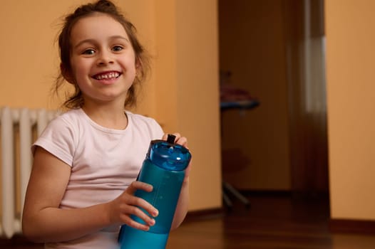 Portrait of 5-6 years old positive sporty child girl holding bottle of water, smiling a toothy smile looking at camera