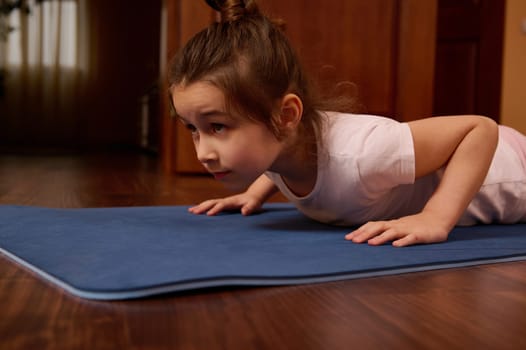 Close-up Caucasian 5-6 years old child, beautiful little girl doing push up on fitness mat indoors