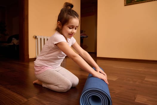 Full length lovely preschooler child girl sitting barefoot on a wooden floor and folding yoga mat after fitness at home