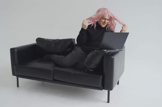 A man in a pink wig, sits on a sofa, communicates via video link on a laptop.