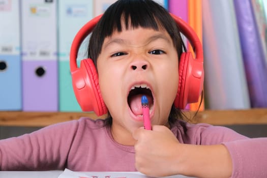 Cheerful little girl in headphones singing and drawing with crayons on paper sitting at table in her room at home. Creativity and development of fine motor skills.