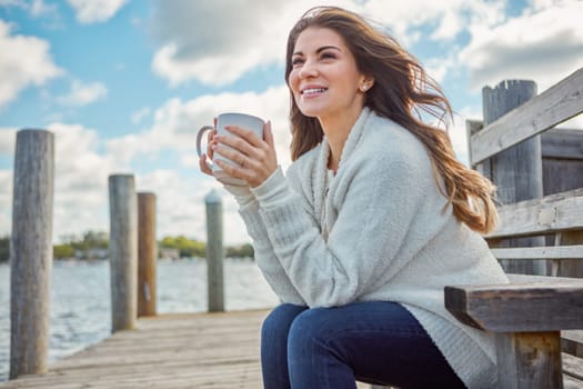 Im grateful for the simple joys of life. a beautiful young woman enjoying a warm beverage while relaxing on a bench at a lake.