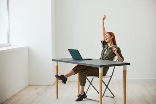 a very happy woman sits at a laptop and rejoices at the completed tasks raising her hands up