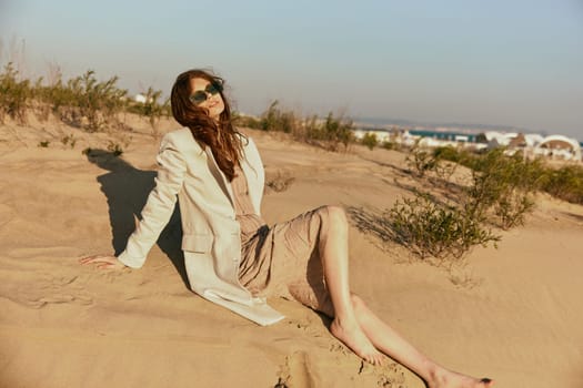 a woman in light clothes and black sunglasses sits on the sand in windy weather. High quality photo