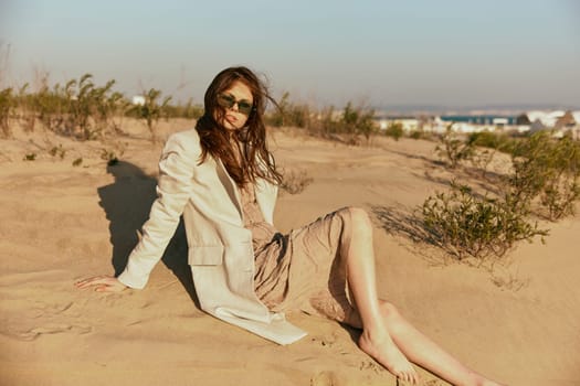 a woman in light clothes and black sunglasses sits on the sand in windy weather. High quality photo