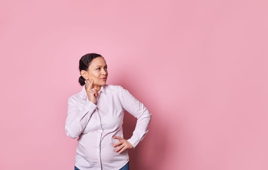 Emotional portrait of a pensive pregnant woman reasoning and looking aside at copy ad space, isolated on pink background