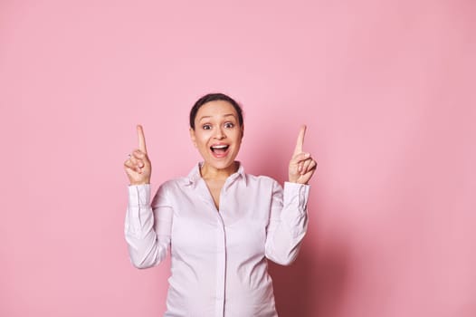 Amazed pregnant woman pointing fingers up, posing with open mouth, expressing positive emotions over pink background