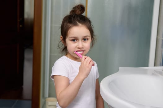 Beautiful preschooler little girl smiling at camera while brushing teeth, standing by sink in the home bathroom.