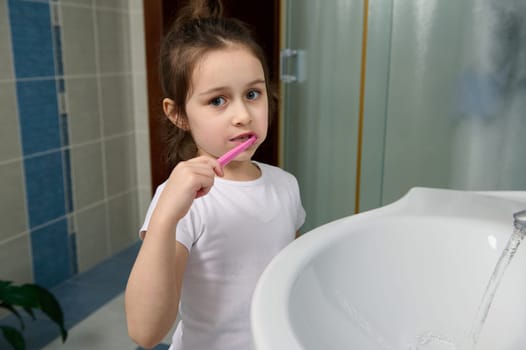 Dental care and oral hygiene for healthy white baby teeth. Adorable little child girl brushing teeth, looking at camera.