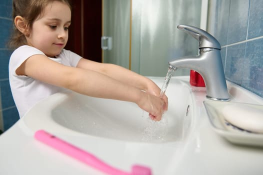 Little kid girl washing hands under tap with running water at home bathroom. Clean hands, hygiene and sanitary concept