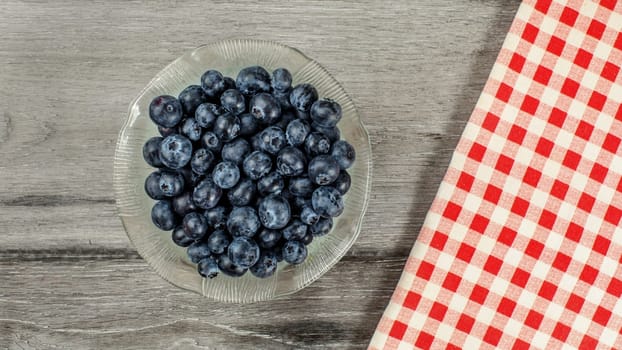 Tabletop view, small glass bowl of blueberries, red checkered gingham tablecloth next to it on gray wood desk.