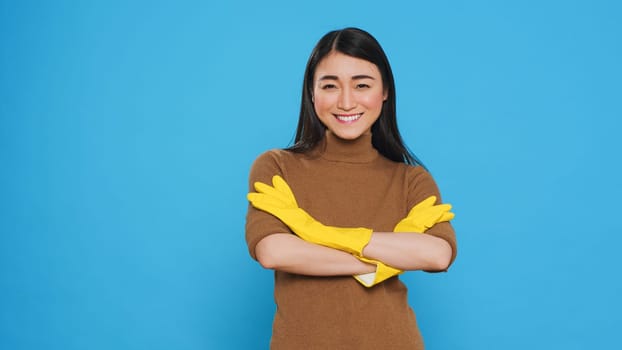 Smiling cleaning lady wearing yellow rubber gloves standing with arm crossed in studio