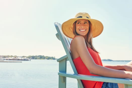 Its a hat wearing kinda day. a cheerful young woman wearing a hat while being seated on a chair next to a lake outside in the sun.