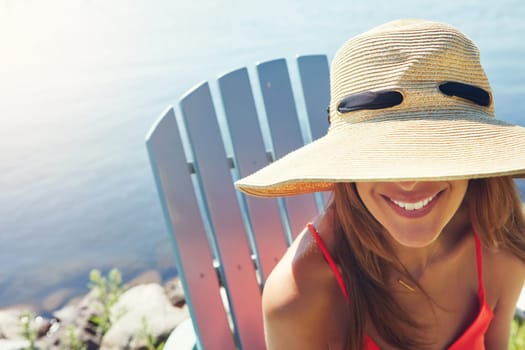 Make sure to wear a hat when the sun is out. a cheerful young woman wearing a hat while being seated on a chair next to a lake outside in the sun.