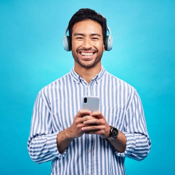 Music, headphones and happy man with phone in studio for streaming, audio and subscription against blue background. Radio, smile and face of asian male online for podcast, track and internet app