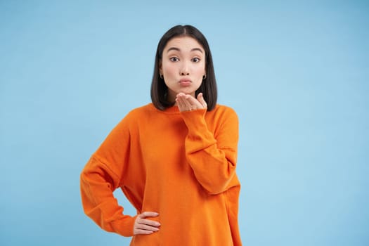Cute young woman puckers her lips and blows air kiss at camera with coquettish face expression, stands over blue studio background