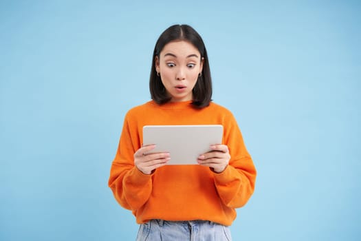 Amazed korean woman with digital tablet, looks surprised and excited, standing over blue background