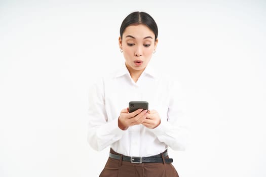 Portrait of asian businesswoman looks at smartphone with surprised face, amazing news on mobile phone message, standing over white background