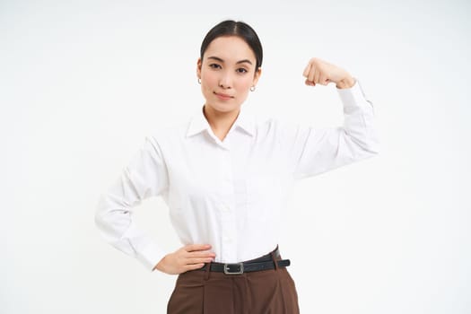 Confident asian businesswoman, flexing biceps, shows strong muscle with serious and self-assured face, white background