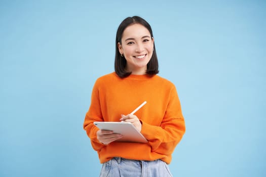 Portrait of young Chinese woman, teacher or student with digital tablet and pencil, writing, taking notes, doing her homework, standing over blue background