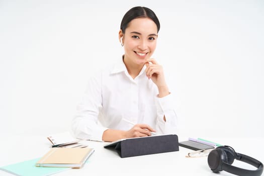 Smiling asian woman sits in her office, works in digital tablet, has headphones, smartphone and work documents on tablet, white background