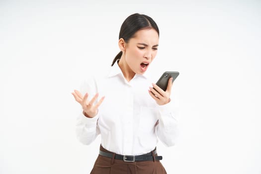 Angry asian businesswoman shouts at her mobile phone with frustrated face expression, stands isolated on white background