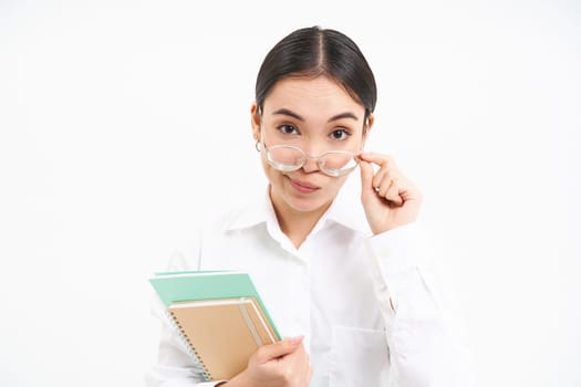 Portrait of korean female teacher, looks under glasses with skeptical face expression, holding notebooks, white background