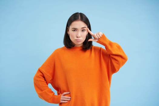 Are you crazy. Young skeptical asian woman holds finger near head, disappointed by someones behaviour, stands over blue background