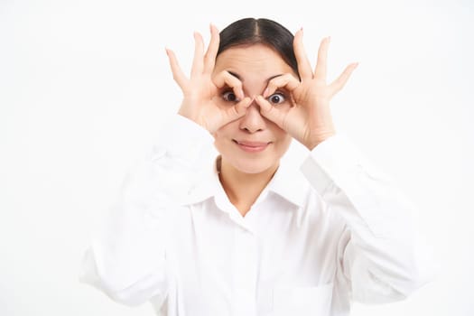 Business people. Happy asian girl looks through finger glasses, stares with surprised face, stands isolated on white background