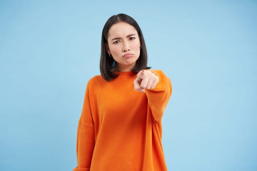 Angry asian woman pointing at camera with blame, blaming you, accusation gesture, standing over blue background