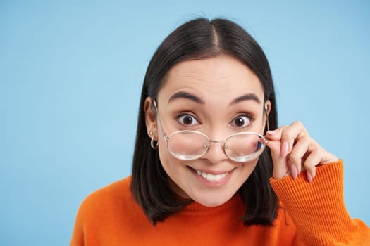 Portrait of brunette japanese woman in glasses, smiling and looking with happy face at camera, tries prescribed eyewear, blue background