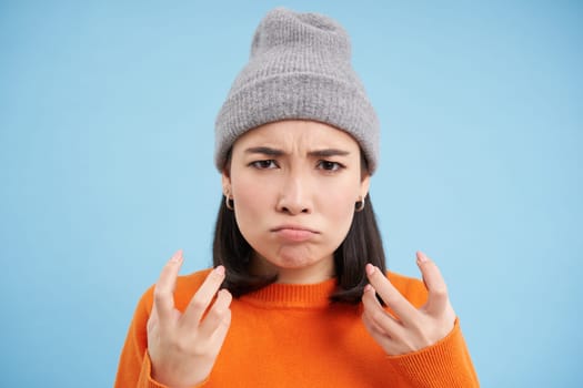 Close up portrait of annoyed asian girl, shakes hands and sulks, looks angry and irritated, stands over blue background